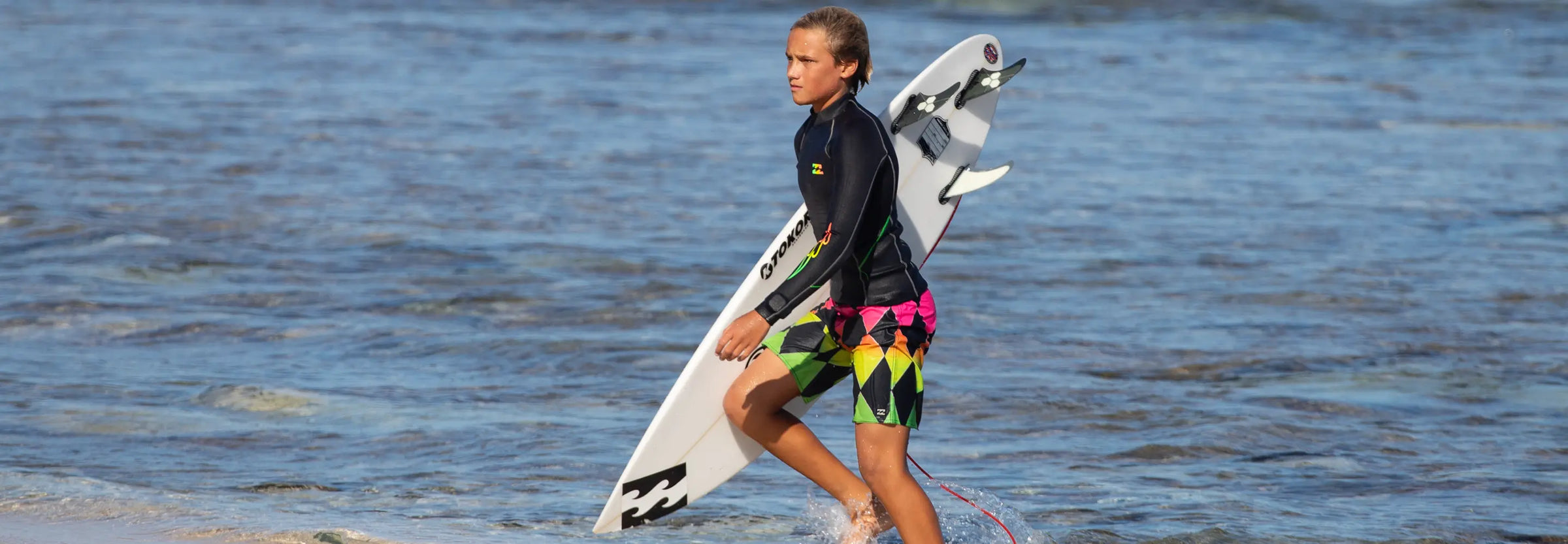 Merrik Mochkatel walking on beach with HIC surfboard in Billabong board shorts and wetsuit