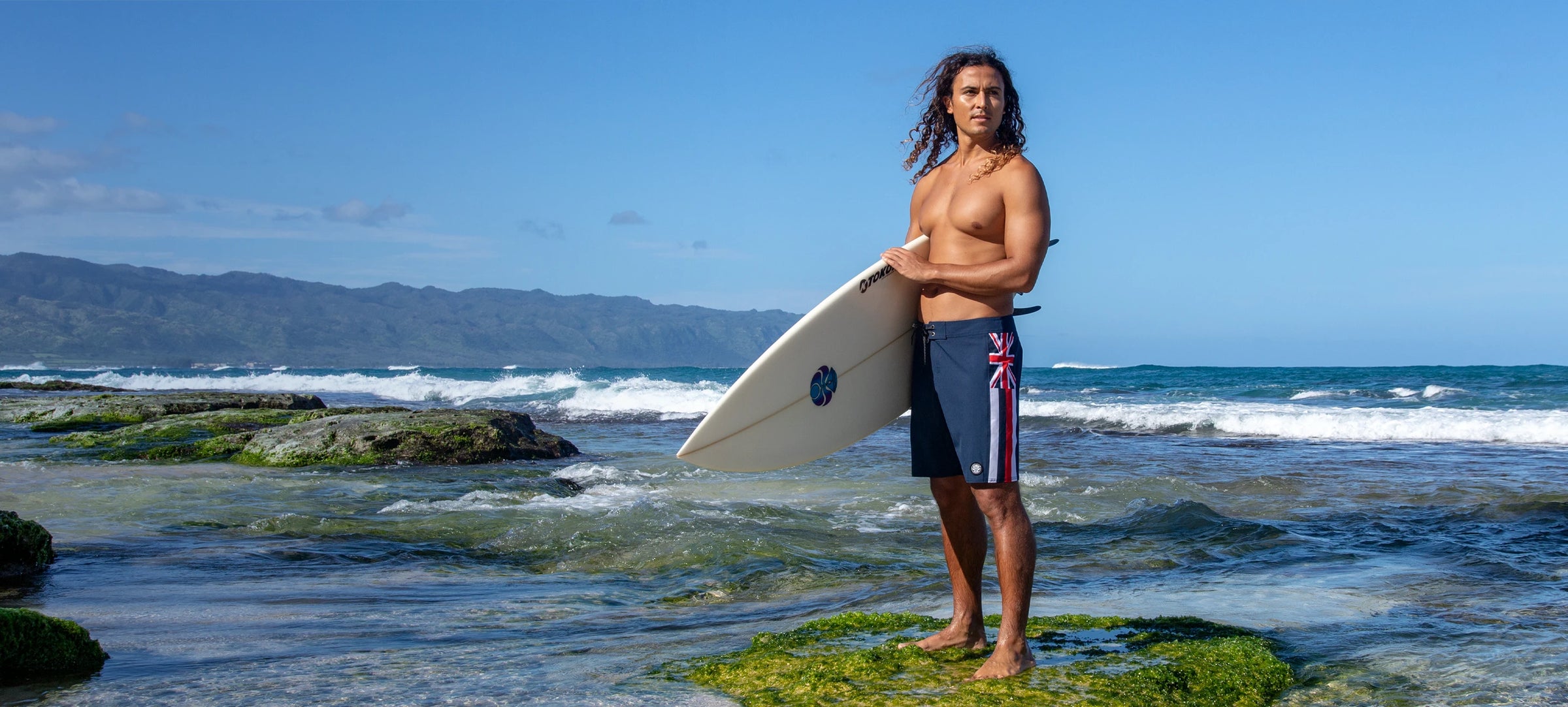 Tiago holding a surfboard on a rocky beach with ocean and mountains in the background