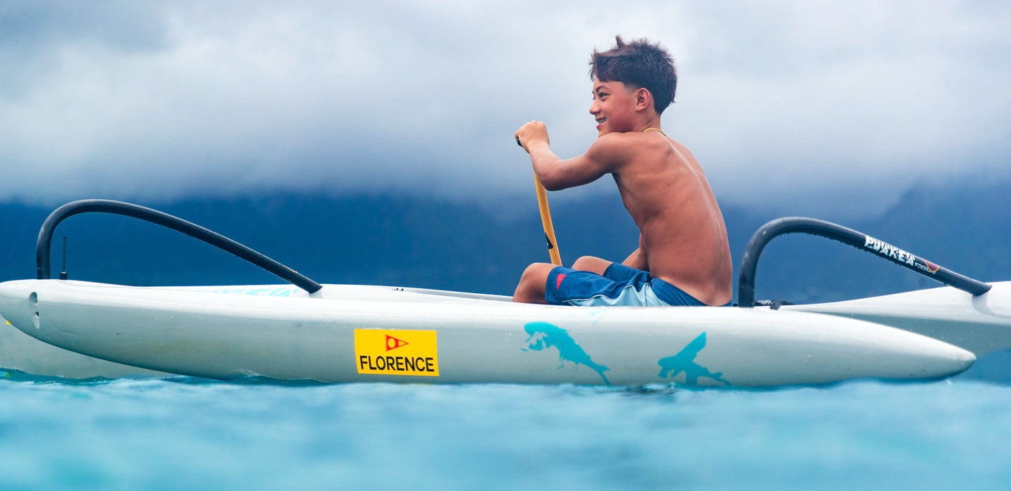 Aukai Tanaka paddling a one-man outrigger canoe on Kailua Bay under a cloudy sky.