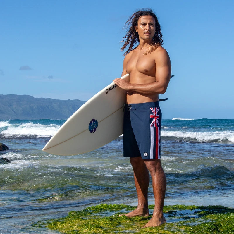Tiago holding a surfboard on a rocky beach with ocean and mountains in the background