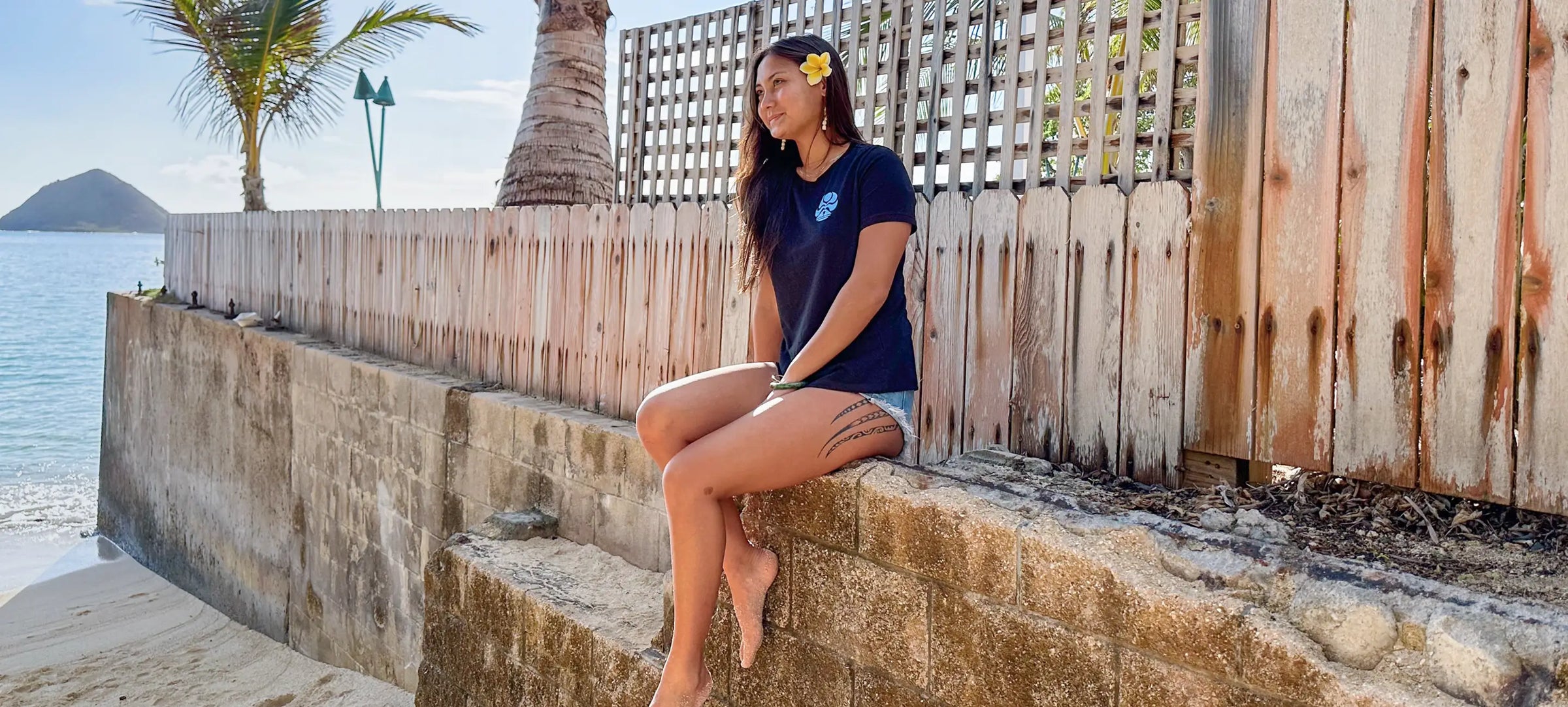 A girl sits on a wall along Lanikai Beach wearing an HIC T-shirt and shorts